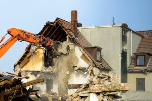 An excavator using traditional demolition methods to tear down a house, creating a large amount of debris and dust. The image contrasts with green demolitions, which focus on environmentally-friendly and controlled dismantling techniques.