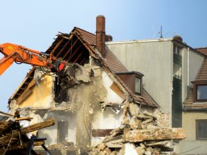 Demolition procedure using heavy machinery to tear down a house, with debris and exposed brick walls visible.