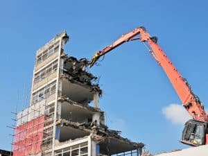 A large excavator demolishes a multi-storey building using a hydraulic arm, showcasing a common demolition method.