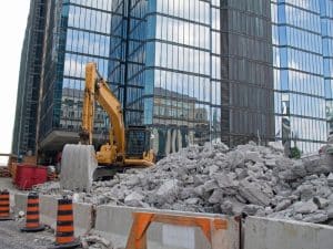 Excavator breaking concrete rubble near modern glass building with focus on effective demolition dust suppression methods.