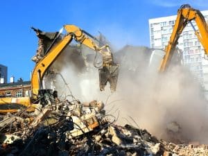 Two yellow excavators demolish a dusty building rubble pile, potentially containing hazardous building materials.