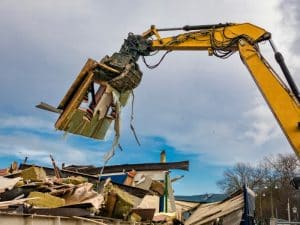 A yellow excavator arm lifts debris containing potential hazardous building materials from a demolished structure against a cloudy sky.