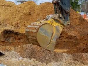A yellow excavator bucket digs into a pile of brown earth, indicating excavation work needed.