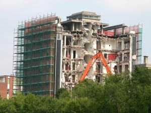 An excavator demolishing a mid-rise apartment block with scaffolding on one side, showing small building demolition in progress.