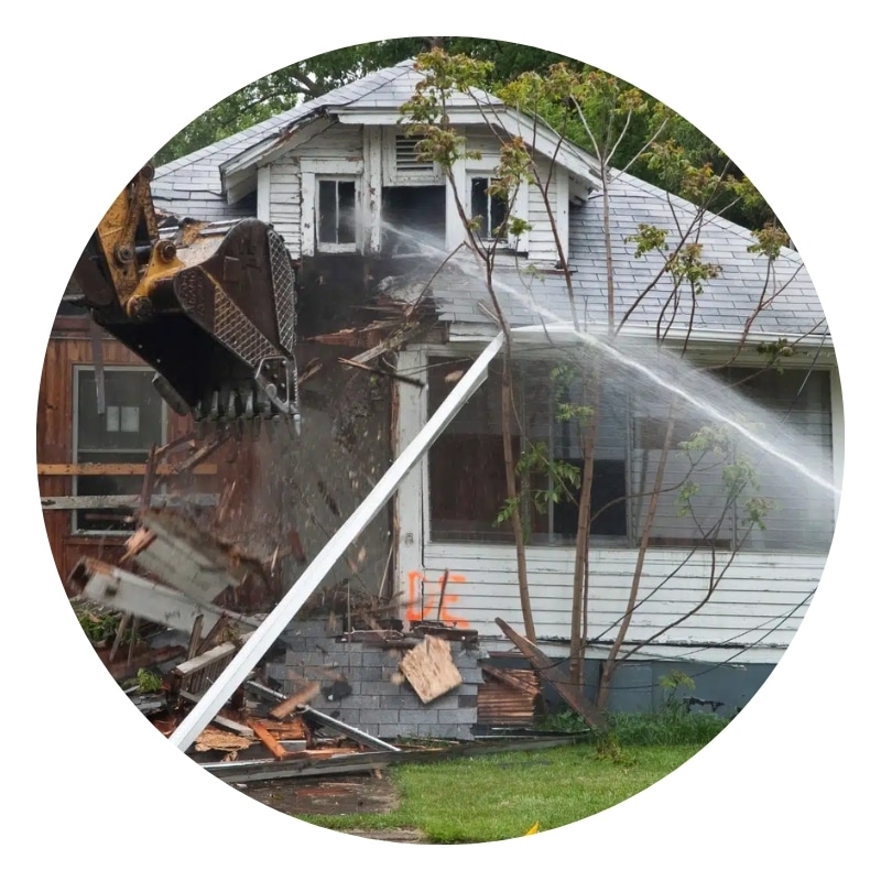 Excavator tearing down house structure with water spray control during residential demolition process.
